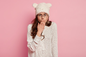 Wall Mural - Shocked overwhelmed woman wears round spectacles, covers mouth with hand, being in stupor from shocking news, isolated over pink background. Amazed girl expresses surprisement and disbelief.