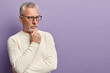 Pensive pensioner holds chin, being deep in thoughts, tries find right solution, wears optical glasses and sweater, isolated over purple background with copy space for your advertisement or text