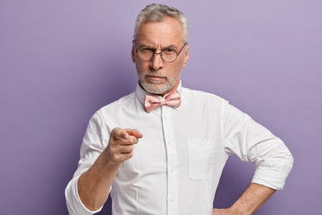 Wall Mural - I want to listen exactly your point of view. Handsome elderly man with thick beard, points at camera with fore finger, has serious expression, dressed in elegant shirt with bowtie, isolated on purple