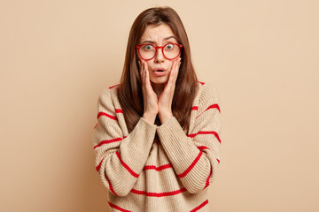 Wall Mural - Worried dark haired lady has stupefied expression, touches cheeks and looks with fright at camera, wears optical glasses for vision correction, loose sweater, isolated over beige background.