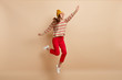 Isolated shot of overjoyed carefree girl jumps high, spreads hands, smiles happily, looks upwards, wears hat with pompon, sweater and red trousers, photographed in motion over beige studio wall