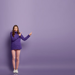 Sticker - Studio shot of astonished female shop assistant in casual winter jumper and rosy trousers, points with both index fingers at free space, stands against purple background. Look in this direction