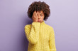 Tired young dark skinned woman purses lips, covers face with palm, closes eyes, feels exhausted, cant continue working, wears yellow knitted jumper, isolated over purple background, wants to sleep.