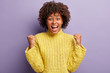 © wayhome.studio  - Overjoyed carefree African American woman raises clenched fists, shows success gesture, triumphs over purple background, wears knitted winter yellow sweater, isolated over purple background.