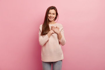 Wall Mural - Impressed friendly pleasant looking woman keeps hands on chest, touched by compliment, smiles positively, wears jumper and jeans, models over pink background, looks at camera with great pleasure