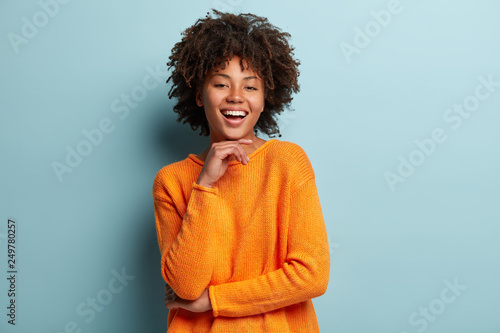 Optimistic Afro American woman with dark skin, curly hair, keeps hands partly crossed, looks at camera with pleasure, wears orange sweater, isolated over blue background. Happy emotions concept.