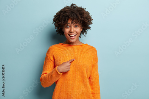 Good looking young African American woman with curly hairstyle, points at herself, wears orange jumper, asks something, has happy expression, isolated over blue background. Do you mean exactly me?