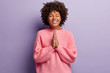 People and spirituality concept. Smiling dark skinned woman with curly bushy hair, keeps hands together, prays for something desirable, isolated over purple background, wears winter sweater.