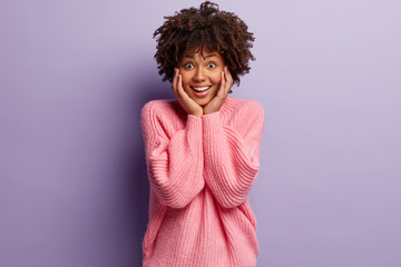Canvas Print - Photo of surprised black woman with Afro hairstyle, keeps hands on cheeks, cant believe in wonderful happy news, wears oversized pink jumper, isolated over purple background. Reaction concept