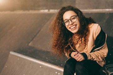 Wall Mural - Smiling mixed race teenage hipster girl sitting at skate park and looking at camera.