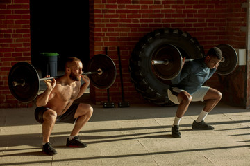 African fitness coach teaches his caucasian male client to lift heavy weights over his head during a pesonal workout session in a gym