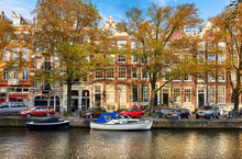 Channel Of Amsterdam City. Netherlands. Motorboats By Banks On Amstel River Among Trees And Traditional Dutch Houses Above Water. Street With Parked Cars. Sunny Spring Day With Blue Sky.