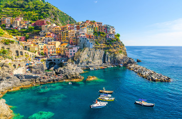 Manarola traditional typical Italian village in National park Cinque Terre, colorful multicolored buildings houses on rock cliff, fishing boats on water, blue sky background, La Spezia, Liguria, Italy