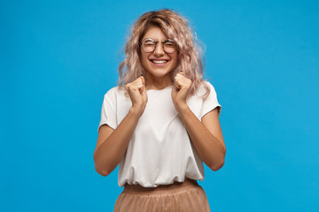 Wall Mural - Success, positive news, joy and happiness concept. Ecstatic excited adorable stylish girl in trendy round glasses smiling broadly and clenching fists, glad to receive present on her birthday