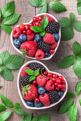 Wall Mural - Berries overhead mix with leaves colorful close up assorted in heart shaped ceramic jar. Raspberry, blueberry, red currant, strawberry on rustic wooden table with green leaves slanted in studio.