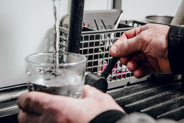 Wall Mural - Pouring water into a glass. Man pours water from a black tap into a transparent glass. The concept of saving water, problems with water, lack of water in various regions of the world.