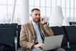 © Margo Kos - Happy businessman working on the laptop and talking on cellphone at the airport waiting lounge. Handsome caucasian businessman at waiting room in airport terminal