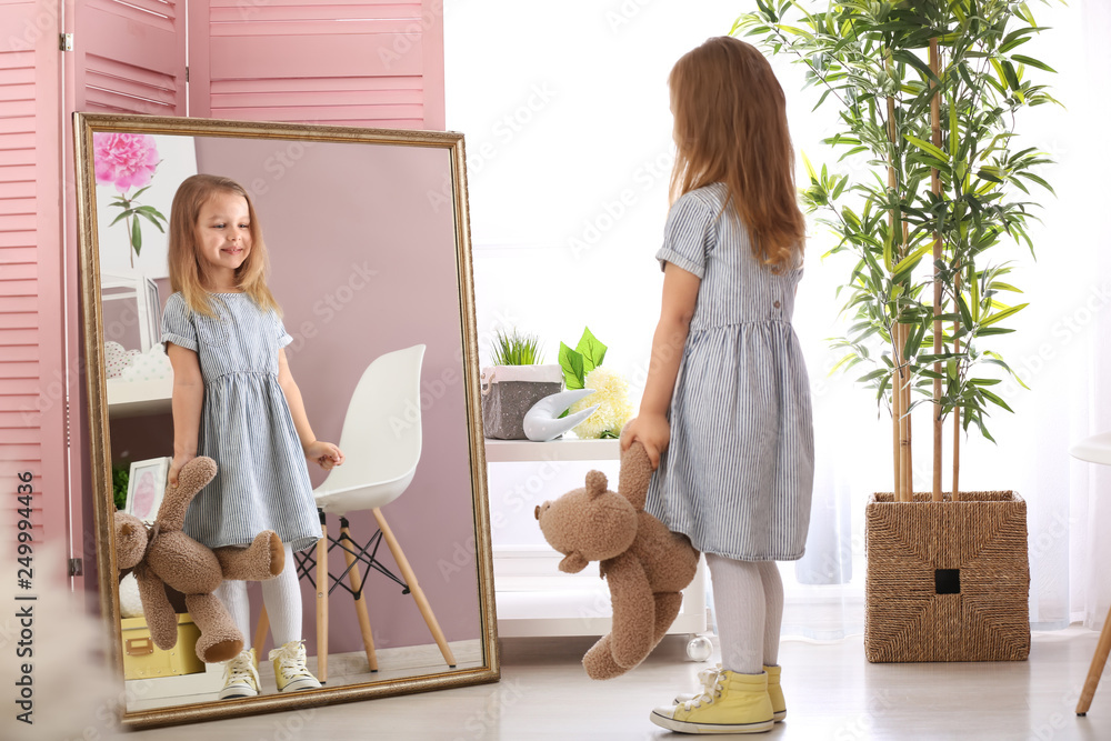 Cute little girl with teddy bear looking in mirror at home - obrazy, fototapety, plakaty 