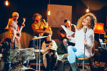 Jazz Band Preparing For The Gig. In Foreground Woman Singing While The Rest Of The Band Playing Bass Guitar, Clavier And Acoustic Guitar. Home Music Studio Interior.