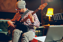 Young Caucasian Bass Guitarist Plauing His Instrument And Holding Cute Little Cat In Lap While Sitting In Home Studio.