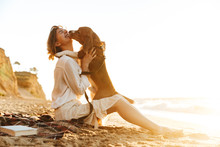 Image Of Pleased Woman 20s Hugging Her Dog, While Sitting On Sand By Seaside