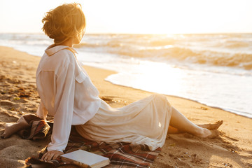 Wall Mural - Image of beautiful woman 20s sitting on sand and looking at sea, while walking along beach