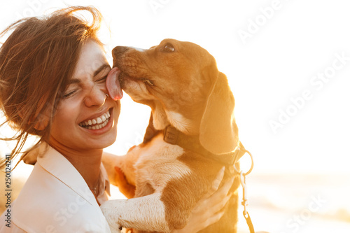 Photographie Image of happy woman 20s hugging her dog, while sitting on sand by seaside