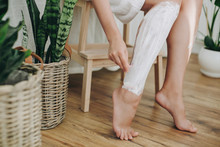Hair Removal Concept, Depilation Process. Young Woman In White Towel Applying Shaving Cream On Her Legs And Holding Holding Plastic Razor In Home Bathroom With Green Plants. Skin Care