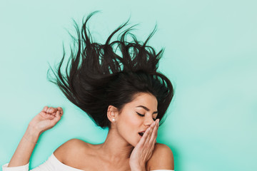 Wall Mural - Beautiful yawning sleepy young woman lies isolated on blue light background. Healthy hair concept.