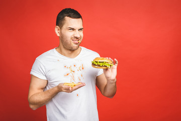 Funny hungry bearded man eating junk food. Excited young man greedily eating hamburgers isolated on red background.