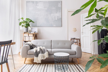 Modern scandianvian decor of living room with design sofa with elegant blanket, coffee table and bookstand on the white wall. Brown wooden parquet. Concept of minimalistic interior.