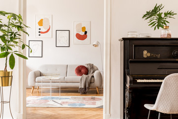 Stylish retro living room with design grey sofa, coffee table, lamp, plants and furniture. Mock up posters frame on the white walls. Minimalistic room with brown wooden parquet and piano. 