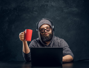 A hacker sits behind a laptop and holding a cup of coffee in a dark office and stares intently at the camera