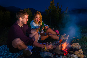 Young couple baking sausages on the campfire and drinking beer in the forest hill in the dusk