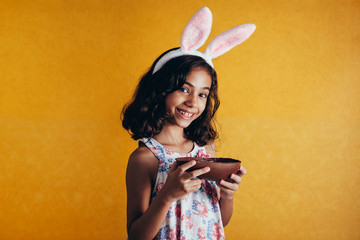 Wall Mural - Cute little child wearing bunny ears on Easter day on color background. Girl eating chocolate easter egg