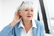 © shurkin_son - Come again please? Attractive mature woman wearing blue jacket and spectacles on her head eavesdropping. Stylish retired female in elegant clothes holding hand at her ear, suffering from hearing loss