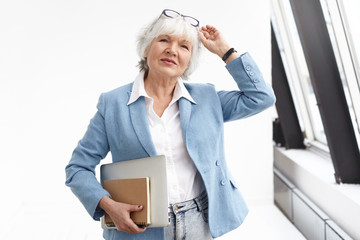 Waist up view of smart classy senior businesswoman wearing stylish blue jacket, jeans and white shirt adjusting glasses on her head, carrying book and laptop, heading to meeting, standing by window