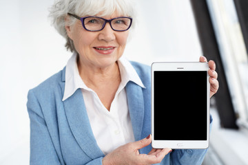 Wall Mural - Electronic devices, gadgets, technology and connection concept. Joyful elegant elderly European gray haired woman in eyewear presenting digital tablet with black display with copyspace for your text