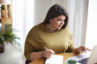© shurkin_son - Indoor shot of overweight plus size beautiful young brunette lady in stylish clothes sitting at desk with open laptop, mug of coffee and writing down information in her diary, studying online