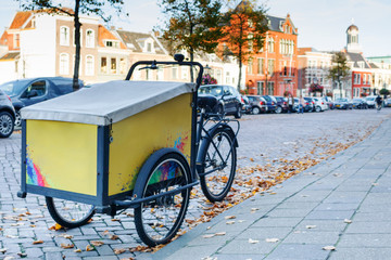 Dutch yellow cargo pedal tricycle parked near sidewalk on the street of Leiden, Netherlands. Historical buildings background. Sunny autumn evening.