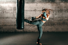 Dedicated Strong Brunette With Ponytail, In Sportswear, Bare Foot And With Boxing Gloves Kicking Sack In Gym.