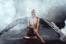 Beautiful Sexy Woman With Black Angel Wings Sitting And Posing On Dark Background
