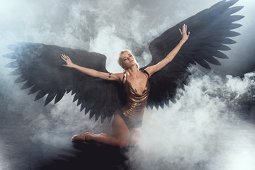 Wall Mural - beautiful sexy woman with black angel wings and outstretched hands sitting and posing on dark smoky background