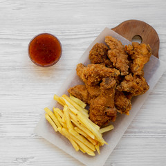 Wall Mural - Fastfood: fried chicken drumsticks, spicy wings, French fries and chicken fingers with sour-sweet sauce over white wooden surface, top view. Flat lay, overhead, from above. Close-up.