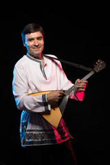 A brunette man in a folk shirt plays a balalaika in scenic blue and red light on a black stage