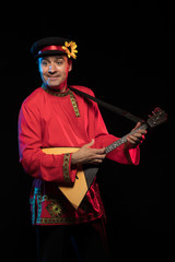 A brunette man in a folk shirt plays a balalaika in scenic blue and red light on a black stage