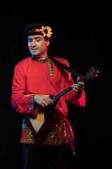 A brunette man in a folk shirt plays a balalaika in scenic blue and red light on a black stage