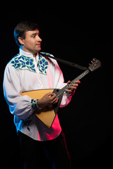 A brunette man in a folk shirt plays a balalaika in scenic blue and red light on a black stage