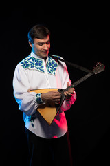 A brunette man in a folk shirt plays a balalaika in scenic blue and red light on a black stage