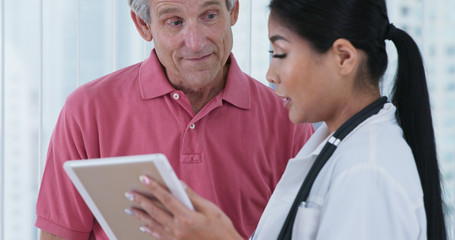 Female primary care physician and her patient discussing test results. Elderly Caucasian male patient talking to Japanese woman doctor while looking at tablet computer together